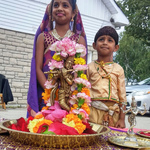 A boy and a girl, dressed as Radha and Krishna, behind a brass Krishna murti
