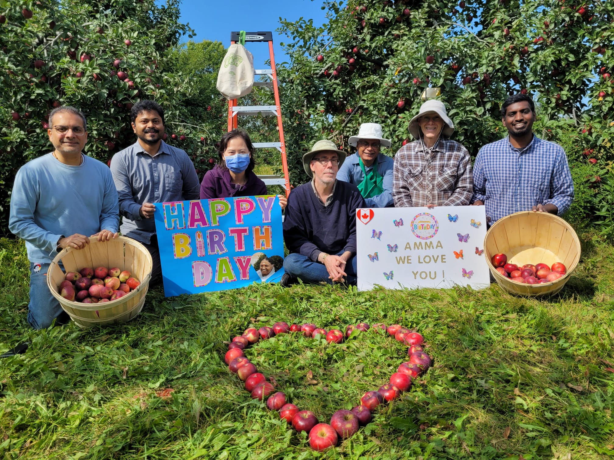 Volunteers in orchard with heart made of apples and signs that read 'Happy Birthday, Amma! We love you!"